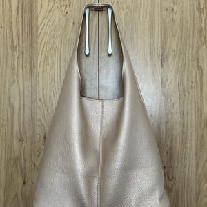 May include: A light pink, leather-textured tote bag with a wide, open top. The bag has a simple, minimalist design with a single vertical seam down the center. The bag is hanging on a silver hook against a wooden background.