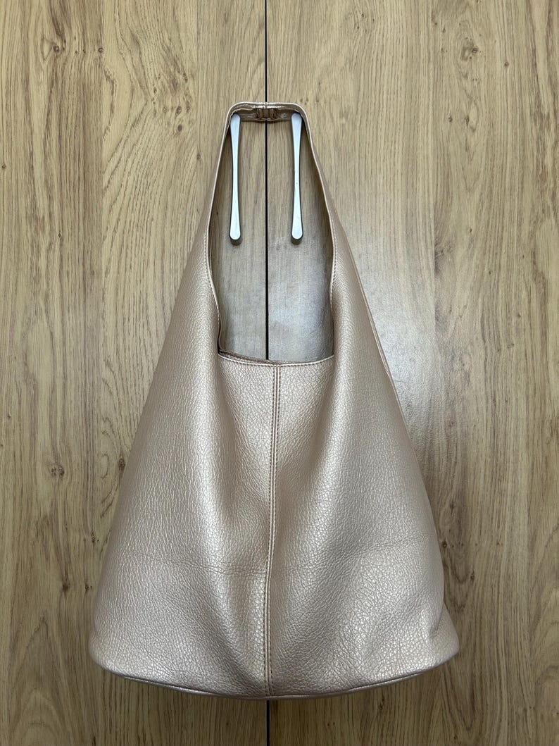 May include: A light pink, leather-textured tote bag with a wide, open top. The bag has a simple, minimalist design with a single vertical seam down the center. The bag is hanging on a silver hook against a wooden background.
