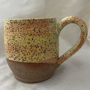 May include: A speckled ceramic mug with a light brown, yellow, and green glaze. The mug has a wide, rounded body and a large handle.