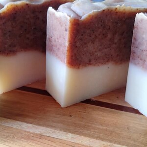 Puede incluir: Primer plano de barras de jabón hechas a mano con un diseño de dos tonos. La mitad inferior es de color blanco cremoso, mientras que la superior es marrón moteado. Limas verdes colocadas sobre una superficie de madera cerca del jabón.