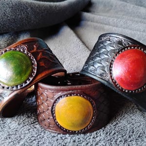 Op de afbeelding: Drie leren armbanden met verschillende ronde inlays in verschillende kleuren. De armbanden zijn bruin, zwart en bruin met groene, rode en gele inlays.