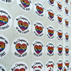 Op de afbeelding: Een vel met ronde, kleurrijke stickers met de tekst "Mrs Chapaneri says My Work is Popping!" en "Mrs Chapaneri says Popping Work!" erop gedrukt. De stickers hebben een regenboog hart met een smiley.
