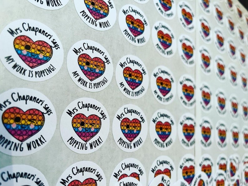 Op de afbeelding: Een vel met ronde, kleurrijke stickers met de tekst "Mrs Chapaneri says My Work is Popping!" en "Mrs Chapaneri says Popping Work!" erop gedrukt. De stickers hebben een regenboog hart met een smiley.