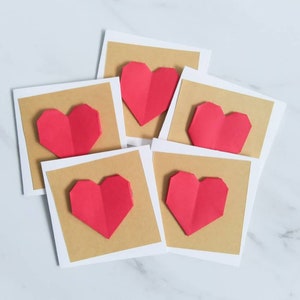 May include: Five handmade cards with white borders and brown interiors. Each card features a red origami heart.