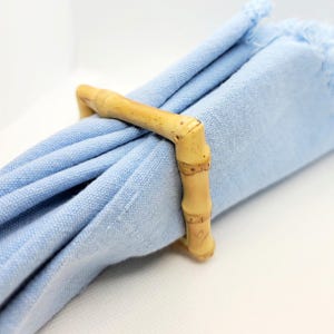 May include: A light blue linen napkin is held by a bamboo napkin ring. The napkin ring is made of natural bamboo and has a cylindrical shape. The napkin is rolled up and has a fringed edge.