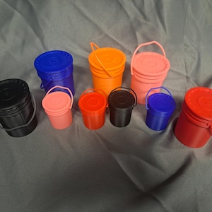 May include: A collection of small, colorful plastic buckets with lids and handles. The buckets are in various colors, including black, blue, orange, pink, and red. The buckets are arranged on a gray surface.