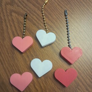May include: A collection of heart-shaped light pull chains in various colors, including pink, white, and red. Each heart is attached to a metal chain with a decorative pull. The chains are arranged on a wooden surface.
