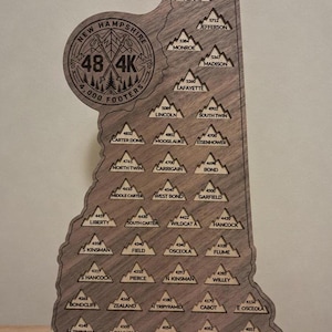 May include: A wooden wall plaque shaped like the state of New Hampshire, featuring engraved mountain icons for the 48 4,000-footers. Each mountain is labeled with its name and elevation in feet, topped by a circular logo reading New Hampshire 48 4K.
