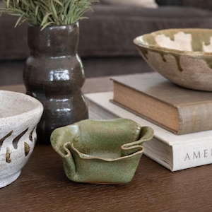 Op de afbeelding: Een verzameling handgemaakte keramische potten op een houten oppervlak. De items omvatten een kleine, gevouwen groene schaal, een donkerbruine vaas met gedroogd groen en twee kommen met aardetinten. Boeken op de achtergrond.