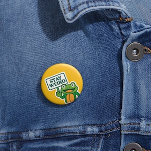 May include: A yellow button pin with the text "STAY WEIRD" and a cartoon frog holding a sign. The frog is green and orange. The pin is on a blue denim jacket.