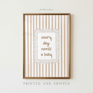 May include: Framed wall art with a gold border. The background features vertical beige and white stripes. The central image has a white scalloped frame with the text "every dog needs a baby" in a handwritten style. The bottom of the frame reads "PRINTED AND SHIPPED".