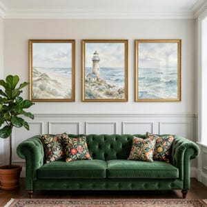May include: A deep green tufted velvet sofa with four patterned throw pillows featuring fruit motifs, positioned beneath three framed watercolor paintings depicting a coastal landscape with a lighthouse, ocean waves, and sandy dunes.