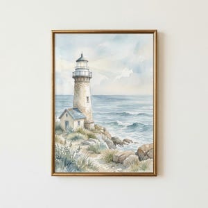 May include: A framed watercolor painting depicting a tall lighthouse standing on a rocky coastline beside a small cottage. The scene features a calm blue ocean with gentle waves, soft clouds in the sky, and green coastal grasses in the foreground.