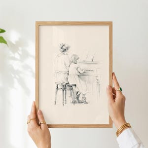 May include: A framed pencil sketch on off-white paper depicting a person sitting on a stool while teaching a child to play the piano. The artwork is enclosed in a light-colored wooden frame with a simple, clean profile and a rectangular shape.