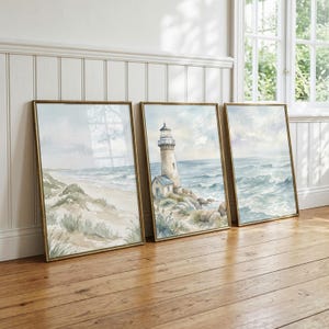 May include: Three framed watercolor prints featuring coastal scenes. The left print shows a sandy beach with dunes, the center print depicts a lighthouse on a rocky shore, and the right print displays ocean waves. Each print has a gold-toned frame.