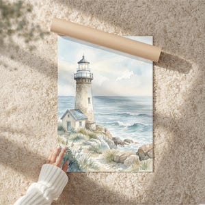 May include: A watercolor print featuring a lighthouse on a rocky coastline with a small adjacent building. The scene depicts a calm sea with soft blue and beige tones, grassy dunes, and a bright light emanating from the top of the lighthouse tower.