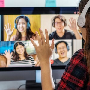 May include: A woman wearing a red and black checked shirt and headphones is waving at a computer screen. The screen shows four people on a video call, all waving back.
