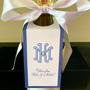 May include: A white wine bottle tag featuring a blue border and a blue monogram with the letters MK. The tag includes the text Cheers from Katie & Michael in blue script. A white satin ribbon is tied around the neck of the glass bottle.
