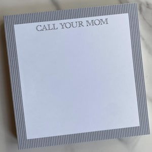 May include: Square notepad with a white interior and a gray striped border. The text "CALL YOUR MOM" is printed in black at the top of the white space. The notepad is on a white and gray marble surface.
