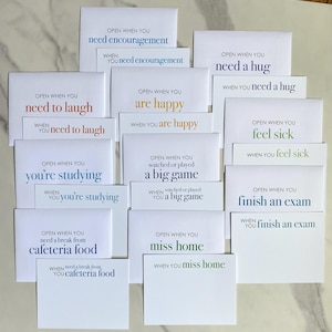 May include: A collection of white envelopes with various phrases printed in different colors. Phrases include "Open When You Need Encouragement," "Need a Hug," "Are Happy," and "Finish an Exam." The envelopes are arranged on a light-colored surface.