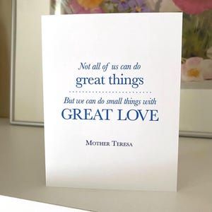 May include: A white greeting card with the quote "Not all of us can do great things, but we can do small things with GREAT LOVE" by Mother Teresa, in blue text. The card is on a white surface.