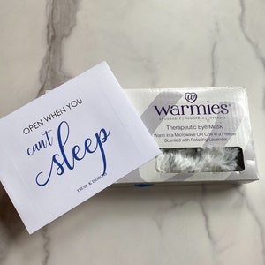 May include: A white eye mask with the text "warmies" in blue. The eye mask is packaged in a white box with blue text that reads "Snuggable Huggable Loveable Therapeutic Eye Mask Warm in a Microwave OR Chill in a Freezer Scented with Relaxing Lavender". A white card with blue text reads "OPEN WHEN YOU can't sleep TRULY K DESIGNS".