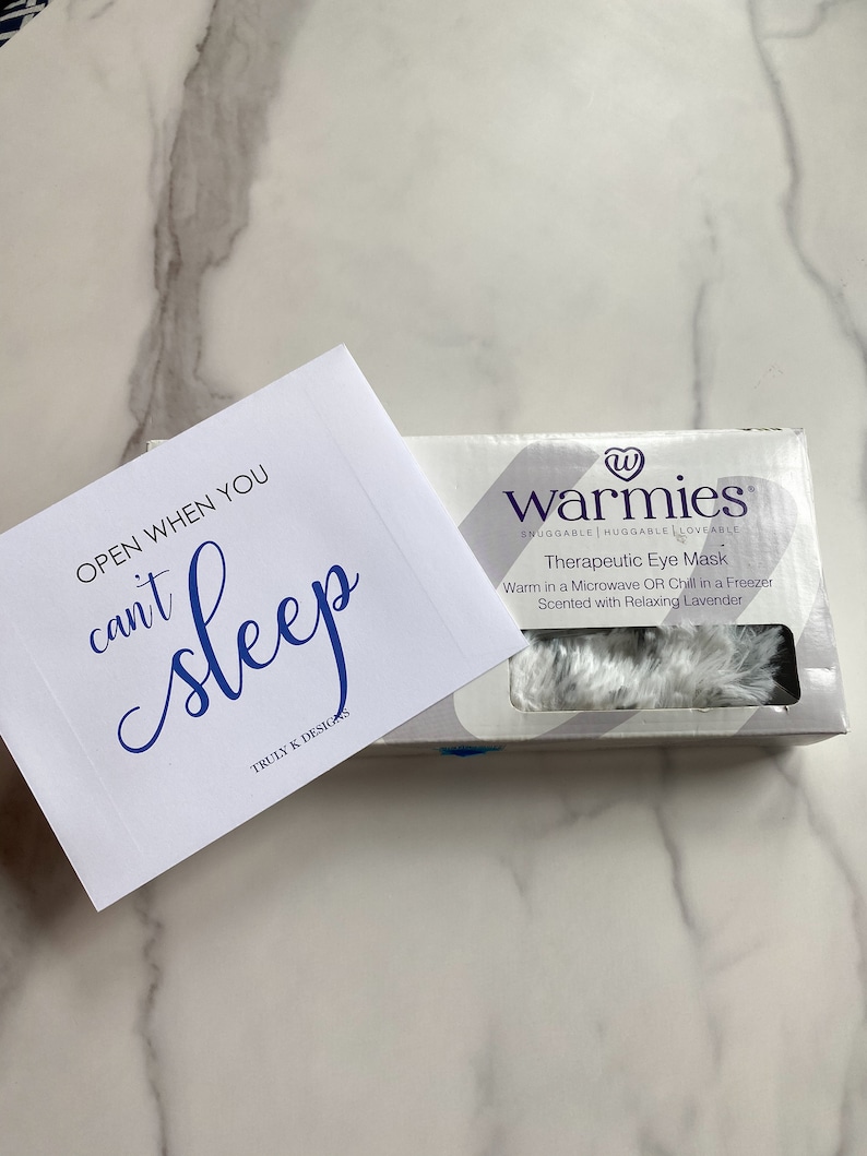 May include: A white eye mask with the text "warmies" in blue. The eye mask is packaged in a white box with blue text that reads "Snuggable Huggable Loveable Therapeutic Eye Mask Warm in a Microwave OR Chill in a Freezer Scented with Relaxing Lavender". A white card with blue text reads "OPEN WHEN YOU can't sleep TRULY K DESIGNS".