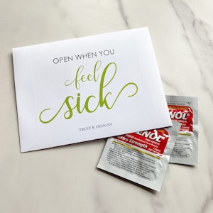 May include: A white card with the text "OPEN WHEN YOU feel sick" in green lettering. The card is sitting on a white surface with two packets of pain reliever medication.