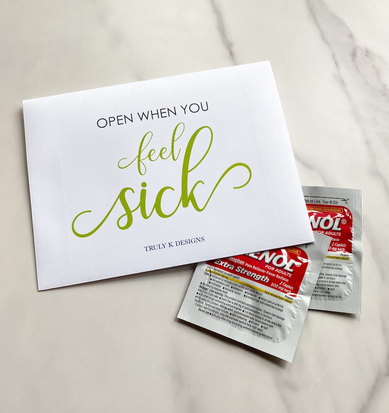 May include: A white card with the text "OPEN WHEN YOU feel sick" in green lettering. The card is sitting on a white surface with two packets of pain reliever medication.