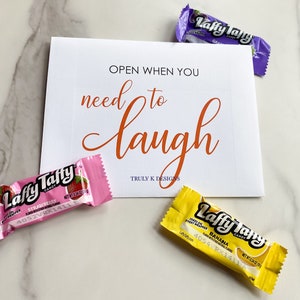 May include: A white card with the text "OPEN WHEN YOU need to laugh" in orange lettering. The card is surrounded by three Laffy Taffy candy wrappers in pink, yellow, and purple. The text "TRULY K DESIGNS" is printed in black at the bottom of the card.