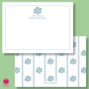 May include: White stationery with a blue floral design and the text "A Note From Sarah Walker." The stationery has a blue border and is paired with a patterned sheet featuring blue hydrangeas and stripes.