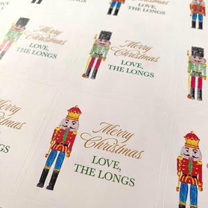 May include: A sheet of rectangular adhesive labels featuring a colourful nutcracker illustration in red, blue, and gold. Each label includes the gold script text Merry Christmas and the green printed text LOVE, THE LONGS in a clean, sans-serif font.