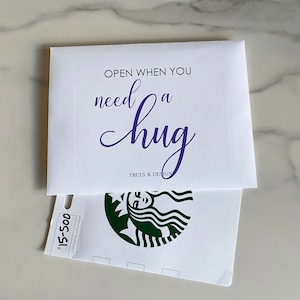 May include: White envelope with the text "OPEN WHEN YOU need a hug" in purple lettering. The envelope is open to reveal a white gift card with a green Starbucks logo and the text "$15-500 card has no value until activated by Starbucks".