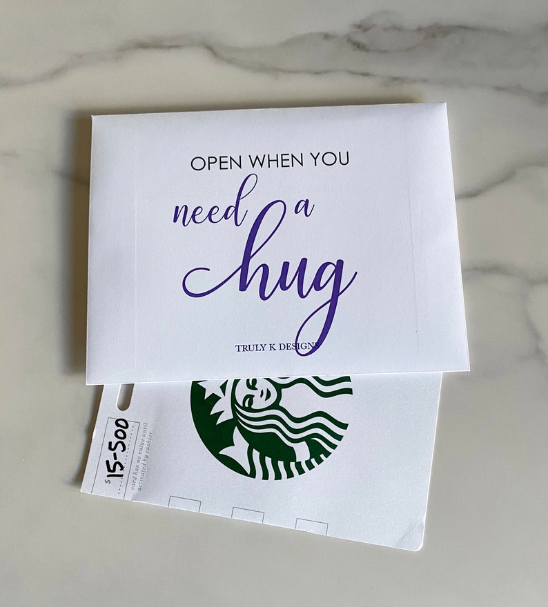 May include: White envelope with the text "OPEN WHEN YOU need a hug" in purple lettering. The envelope is open to reveal a white gift card with a green Starbucks logo and the text "$15-500 card has no value until activated by Starbucks".
