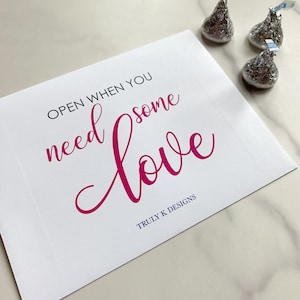 May include: White card with the text "OPEN WHEN YOU NEED SOME LOVE" in pink cursive lettering. The text "TRULY K DESIGNS" is printed in blue below the cursive text. Three silver foil-wrapped chocolate kisses are on a white marble surface.