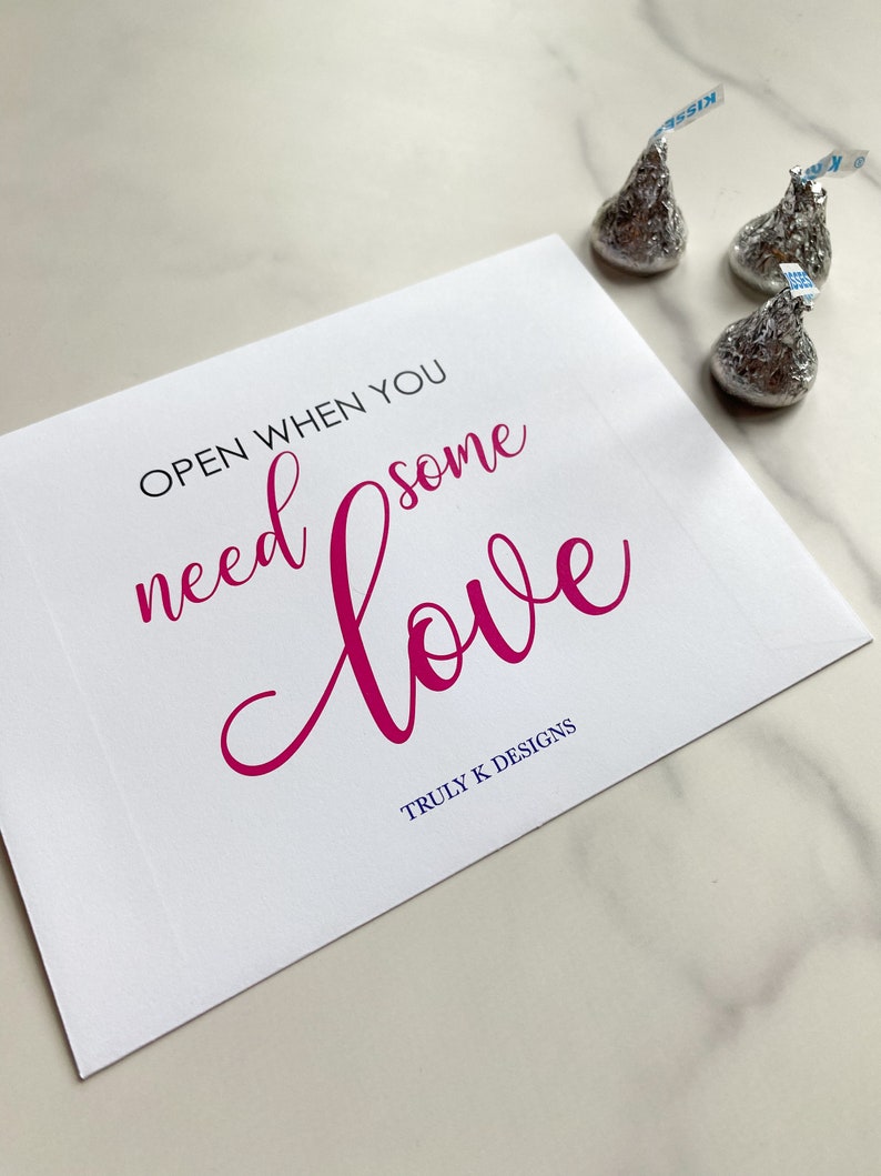 May include: White card with the text "OPEN WHEN YOU NEED SOME LOVE" in pink cursive lettering. The text "TRULY K DESIGNS" is printed in blue below the cursive text. Three silver foil-wrapped chocolate kisses are on a white marble surface.