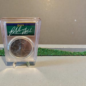May include: A silver coin encased in a clear plastic holder, displayed on a clear stand. The coin features a woman walking, and the holder has a green and blue label with the text "QUACK" and "Celebrity Mint".