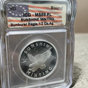 May include: A silver round coin featuring a sunburst eagle design, encapsulated in a clear plastic ICG slab. The label reads ND, Round, ICG - MS69 PL, SUNSHINE MINTING, and Sunburst Eagle, 1/2 Oz.Ag, with an American flag graphic background.