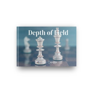 Op de afbeelding: Boekomslag met de titel "Depth of Field" en de woorden "Focused Camera" eronder. De omslag toont schaakstukken op een schaakbord, met de koningin en de koning in focus. De achtergrond is een wazige teal kleur.