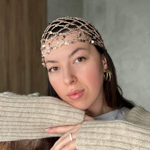 May include: A beige headpiece with a net-like design, adorned with sequins and small beads. The headpiece is worn on the head. The person is wearing gold hoop earrings and a beige sweater.
