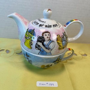 May include: A decorative ceramic teapot and teacup set featuring illustrations from The Wizard of Oz. The teapot has a pink and white design with a rainbow handle. The teacup is blue and white. The set includes the text "Whopper! Better get under cover..."