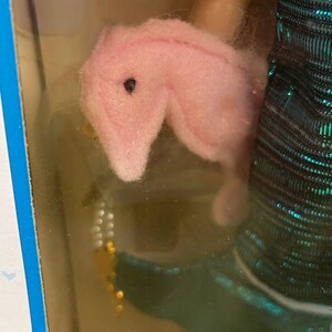 Puede incluir: Un juguete de peluche rosa con forma de caballito de mar con un ojo negro se sostiene cerca de un vestido de mu&ntilde;eca con lentejuelas verde azulado y negro. Tambi&eacute;n es visible un accesorio de cuentas doradas y blancas. El juguete est&aacute; en un paquete de pl&aacute;stico transparente.