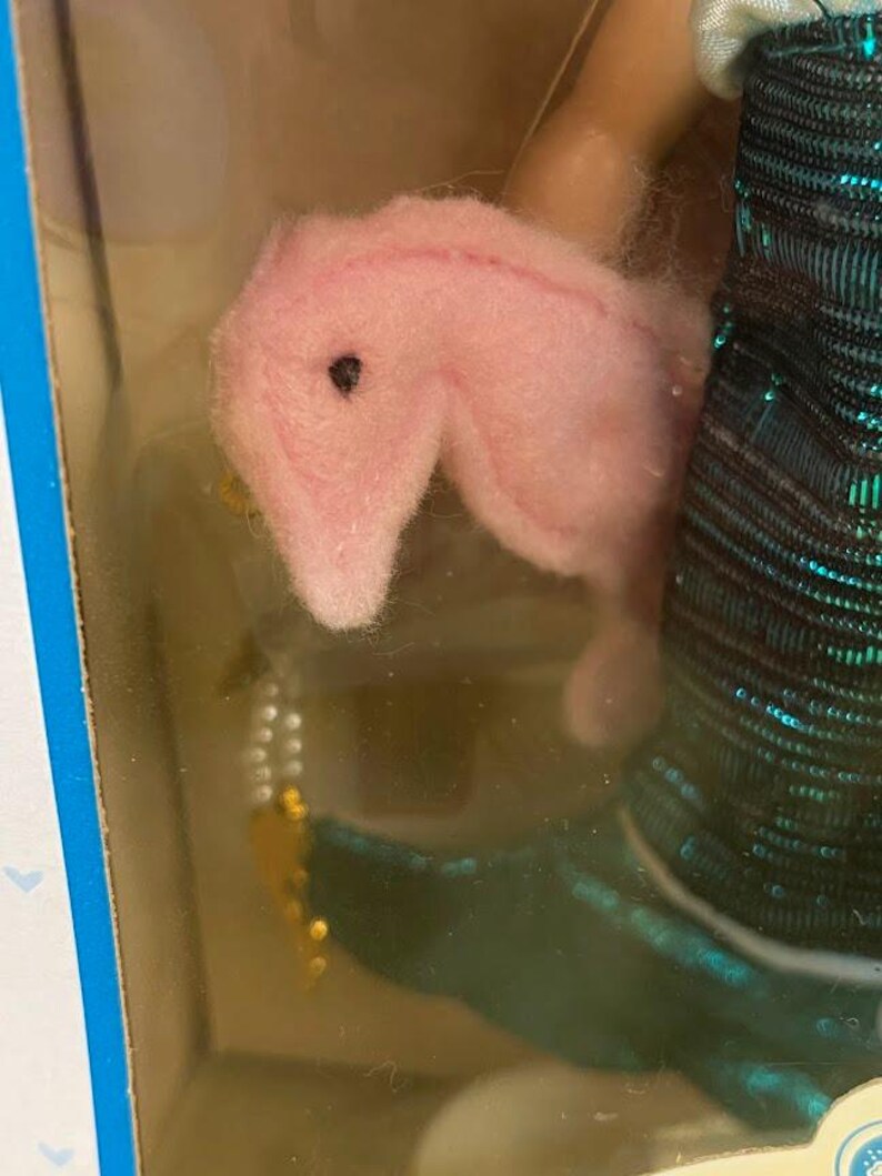 Puede incluir: Un juguete de peluche rosa con forma de caballito de mar con un ojo negro se sostiene cerca de un vestido de mu&ntilde;eca con lentejuelas verde azulado y negro. Tambi&eacute;n es visible un accesorio de cuentas doradas y blancas. El juguete est&aacute; en un paquete de pl&aacute;stico transparente.