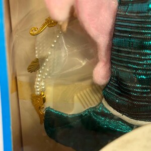 Puede incluir: Una mu&ntilde;eca con una cabeza de peluche rosa de caballito de mar y un atuendo de lentejuelas turquesa. La mu&ntilde;eca sostiene un collar de perlas con dijes dorados de conchas marinas e hipocampos. La mu&ntilde;eca est&aacute; en una caja con ribete azul.