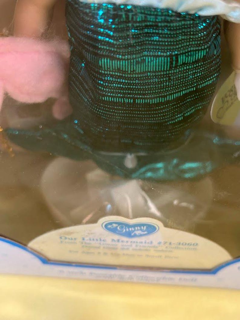 Puede incluir: Una mu&ntilde;eca de sirena en una caja expositora. La mu&ntilde;eca tiene una falda de lentejuelas turquesa y una parte superior blanca. La caja tiene el texto "Ginny" y "Our Little Mermaid #71-3060". Se ve un accesorio rosa y esponjoso.