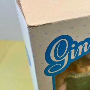 Puede incluir: Primer plano de una caja de mu&ntilde;eca con el nombre "Gin" en fuente azul. La caja es de color rosa claro con peque&ntilde;os patrones de corazones azules. La esquina de la caja est&aacute; ligeramente da&ntilde;ada.