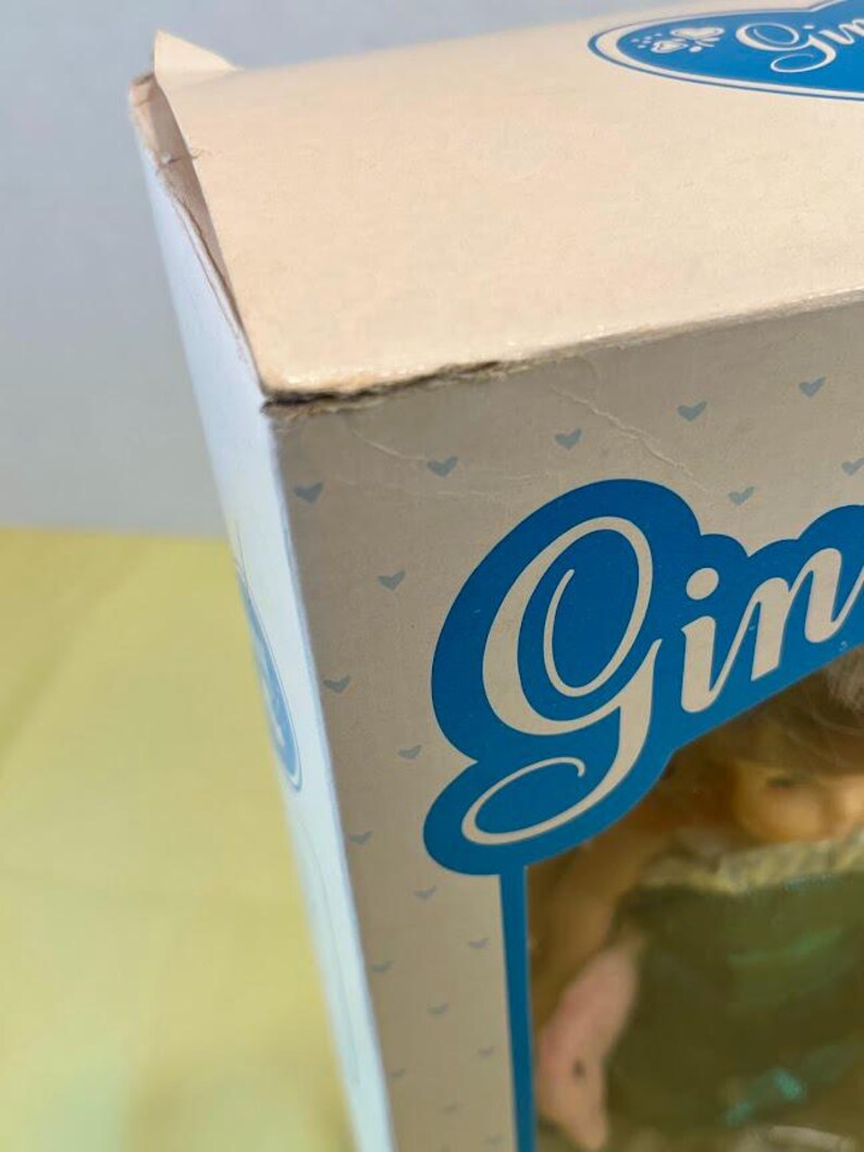 Puede incluir: Primer plano de una caja de mu&ntilde;eca con el nombre "Gin" en fuente azul. La caja es de color rosa claro con peque&ntilde;os patrones de corazones azules. La esquina de la caja est&aacute; ligeramente da&ntilde;ada.