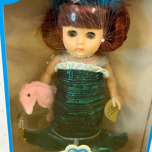 Puede incluir: Una mu&ntilde;eca Ginny vintage, "Our Little Mermaid", en su caja original. La mu&ntilde;eca tiene cabello rojo, un collar de perlas y un vestido de sirena con lentejuelas verdes. Sostiene un accesorio de caballito de mar rosa y lleva un tocado azul y blanco. La caja tiene un dise&ntilde;o azul y blanco.