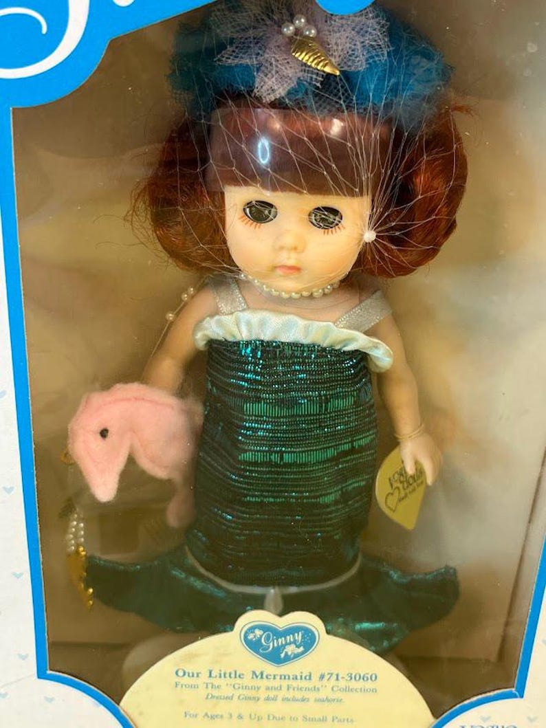 Puede incluir: Una mu&ntilde;eca Ginny vintage, "Our Little Mermaid", en su caja original. La mu&ntilde;eca tiene cabello rojo, un collar de perlas y un vestido de sirena con lentejuelas verdes. Sostiene un accesorio de caballito de mar rosa y lleva un tocado azul y blanco. La caja tiene un dise&ntilde;o azul y blanco.
