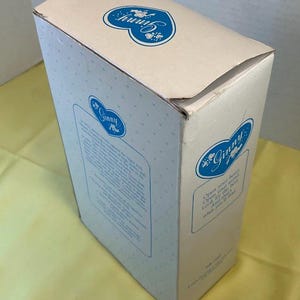 Puede incluir: Una caja de color crema con logotipos azules en forma de coraz&oacute;n floral y el nombre "Ginny". La caja tiene el texto "Abre tu coraz&oacute;n, Abre tu mente, Busca lo mejor, Y lo que encontrar&aacute;s."